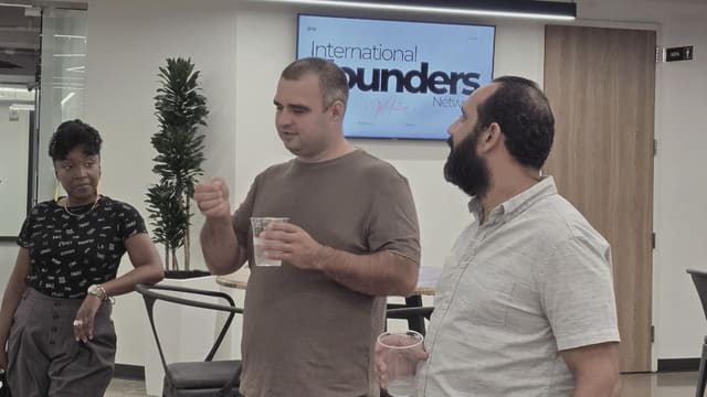 Three people standing together in an open room, the one in the middle speaking with a raised hand, two of them holding drinks, with an IFN meetup slide on the screen behind them.