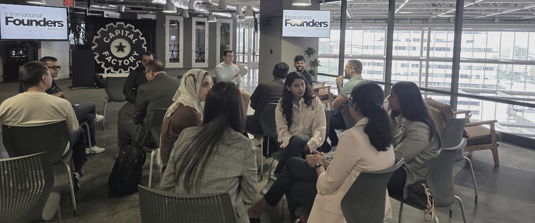 A wide circle of people seated in conversation across a daylit floor, several of them turned toward the camera and one standing at the back mid gesture, with IFN meetup slides on two screens and downtown Austin through a full height glass wall.