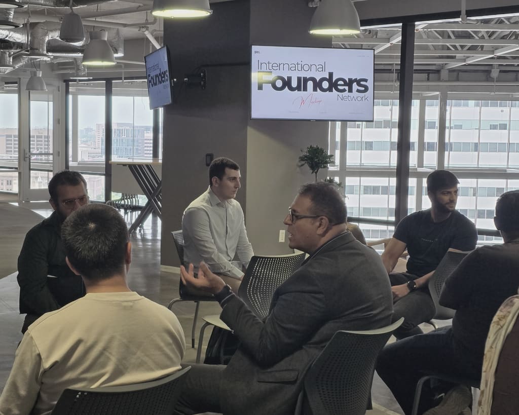 One founder mid gesture in a small seated circle with the others listening, an IFN meetup slide on the screen behind them and the city through the window.